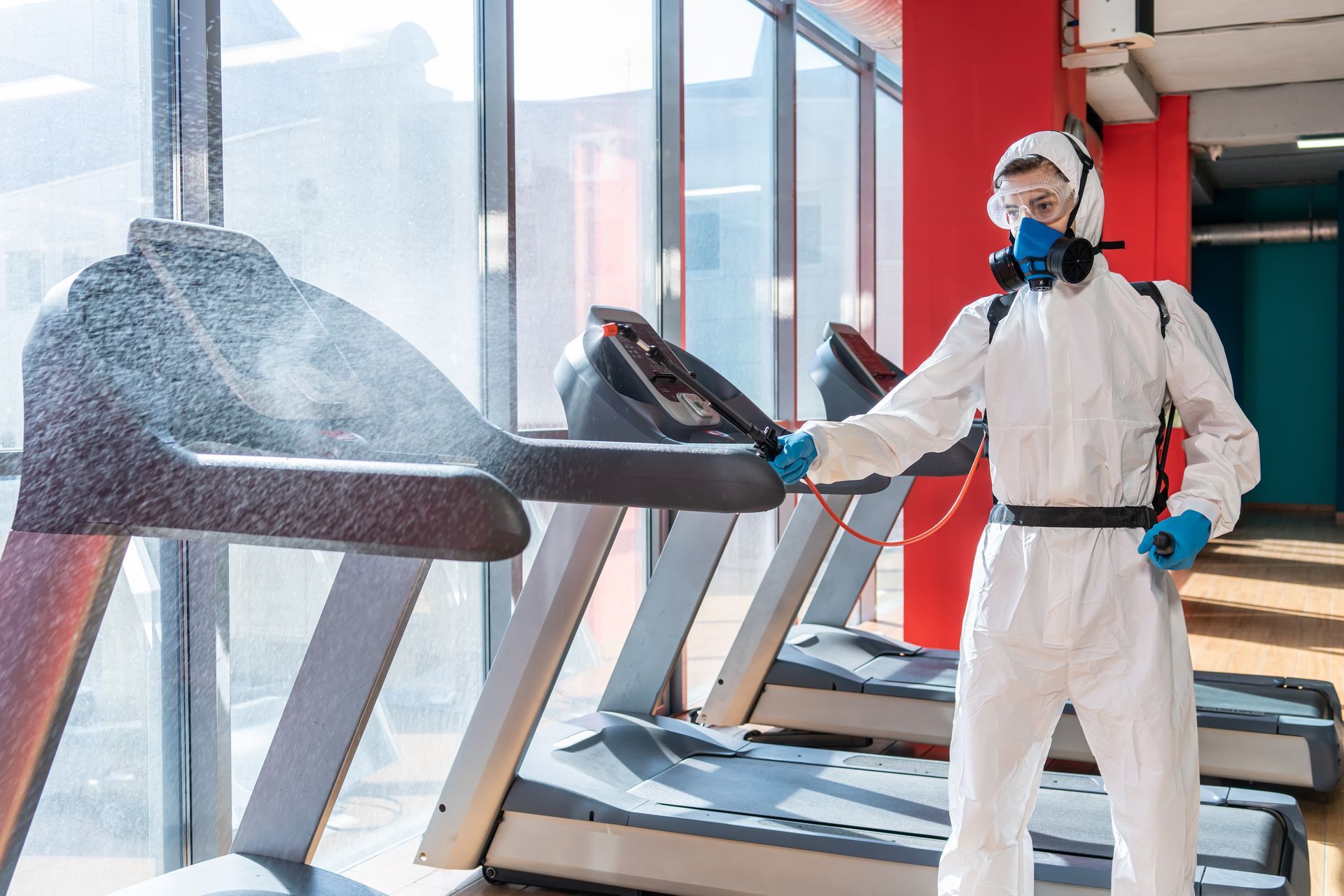 A man in a protective suit is disinfecting treadmills in a gym.