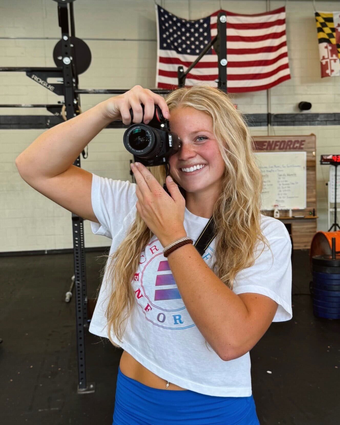 Woman with camera, smiles, holds camera up in gym; American flag behind.