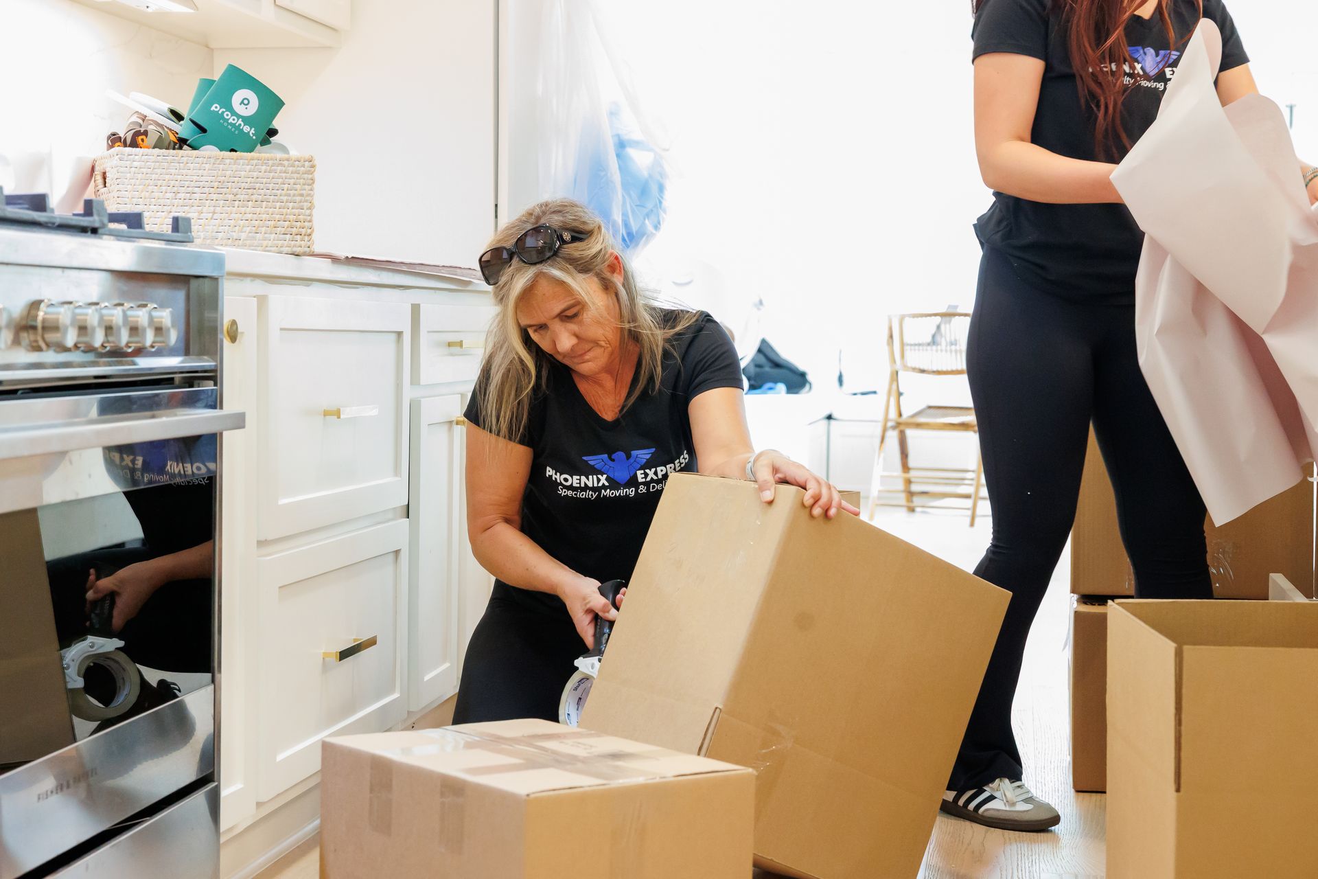 Two people packing boxes in a kitchen with a stove and cabinets. One kneels, cutting tape. Other holds packing paper.