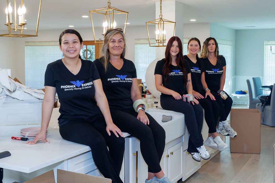 Five people in black shirts and pants sitting on a white counter. Smiling in a room with packed boxes.
