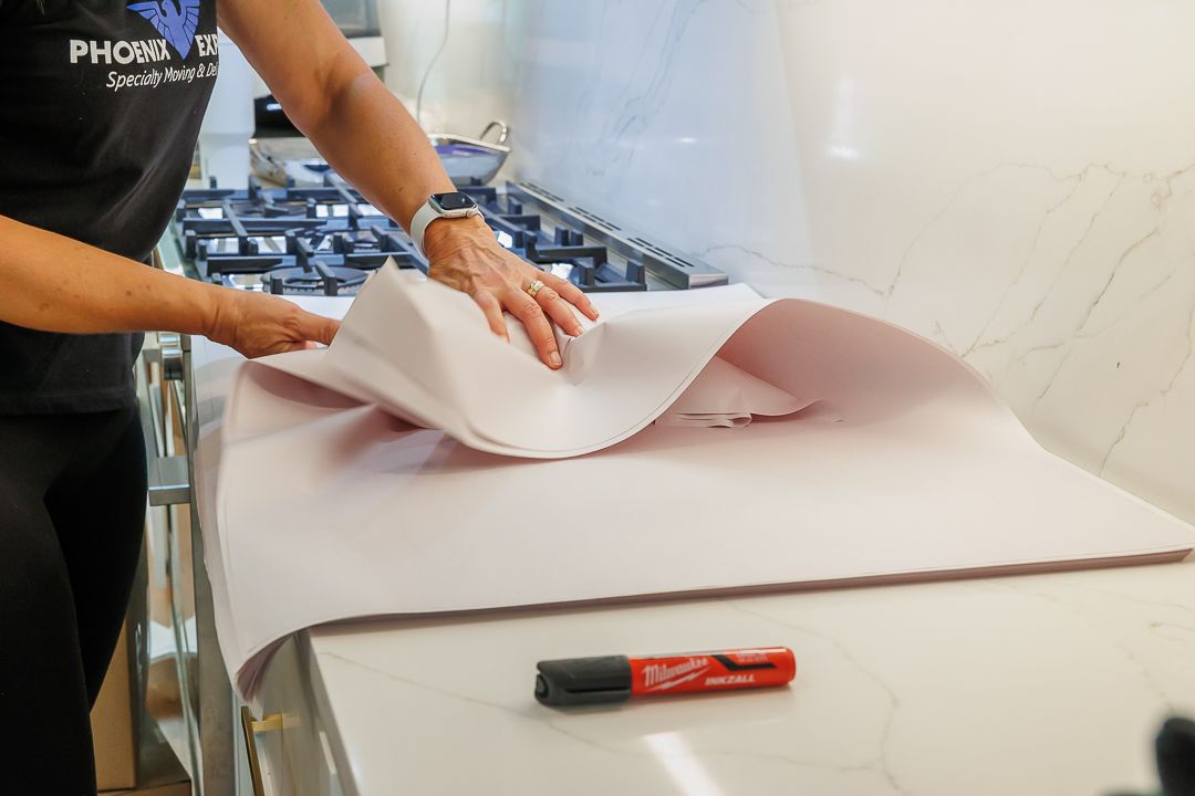 Person unfolding pink paper on a white countertop near a stove. A marker is in the foreground.