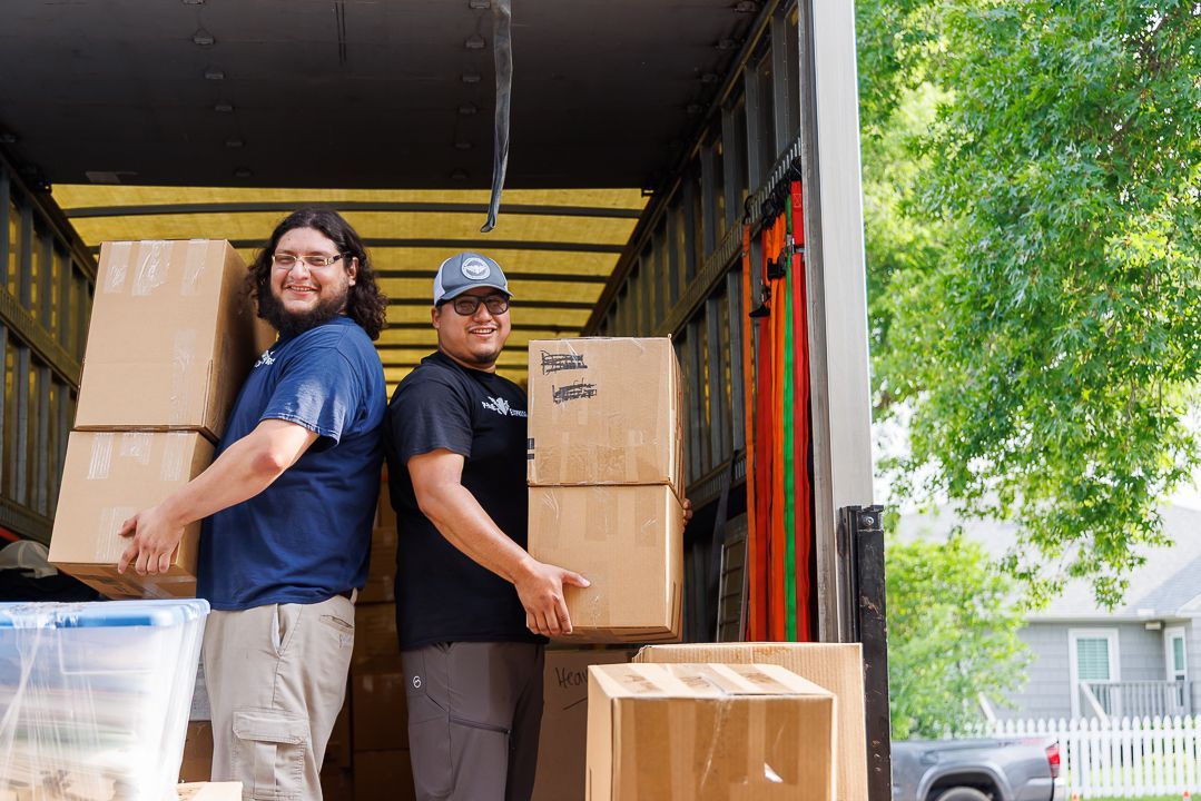Two men load cardboard boxes into a moving truck, smiling. Exterior shot, sunny.