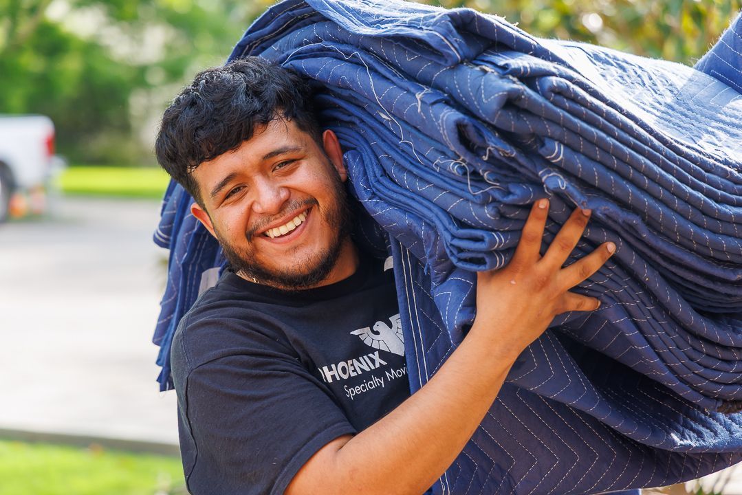 Man carrying stack of blue moving blankets, smiling outdoors.