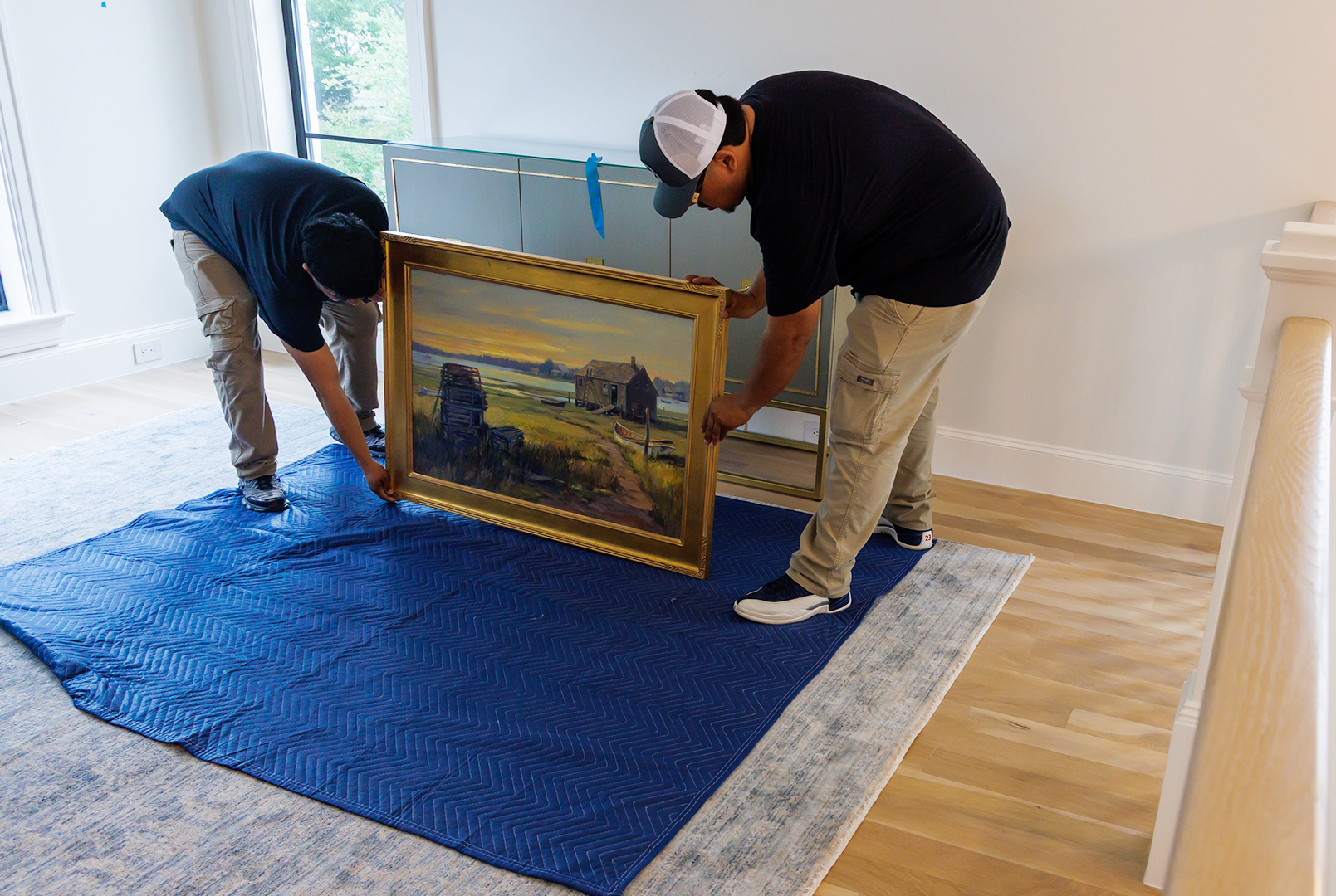 Two movers carefully place a framed painting on a protective blue pad, near a staircase.