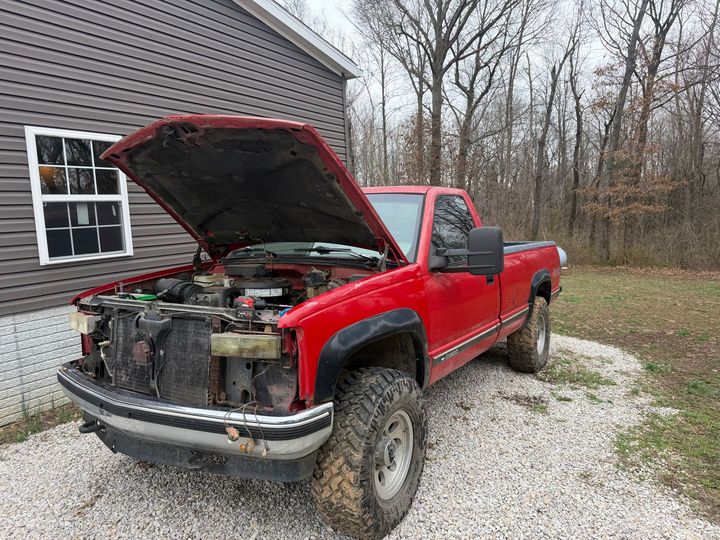 A red pickup truck with its hood open and missing its front grille, parked on a gravel driveway in front of a house.