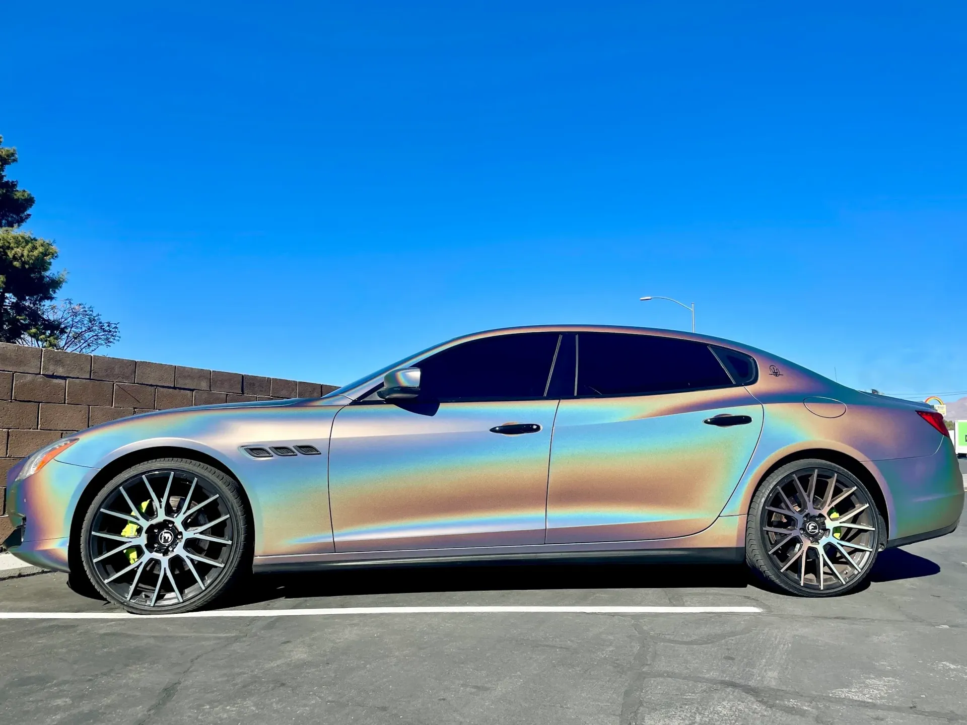 Rainbow-iridescent sedan parked beside a stone wall under a bright blue sky