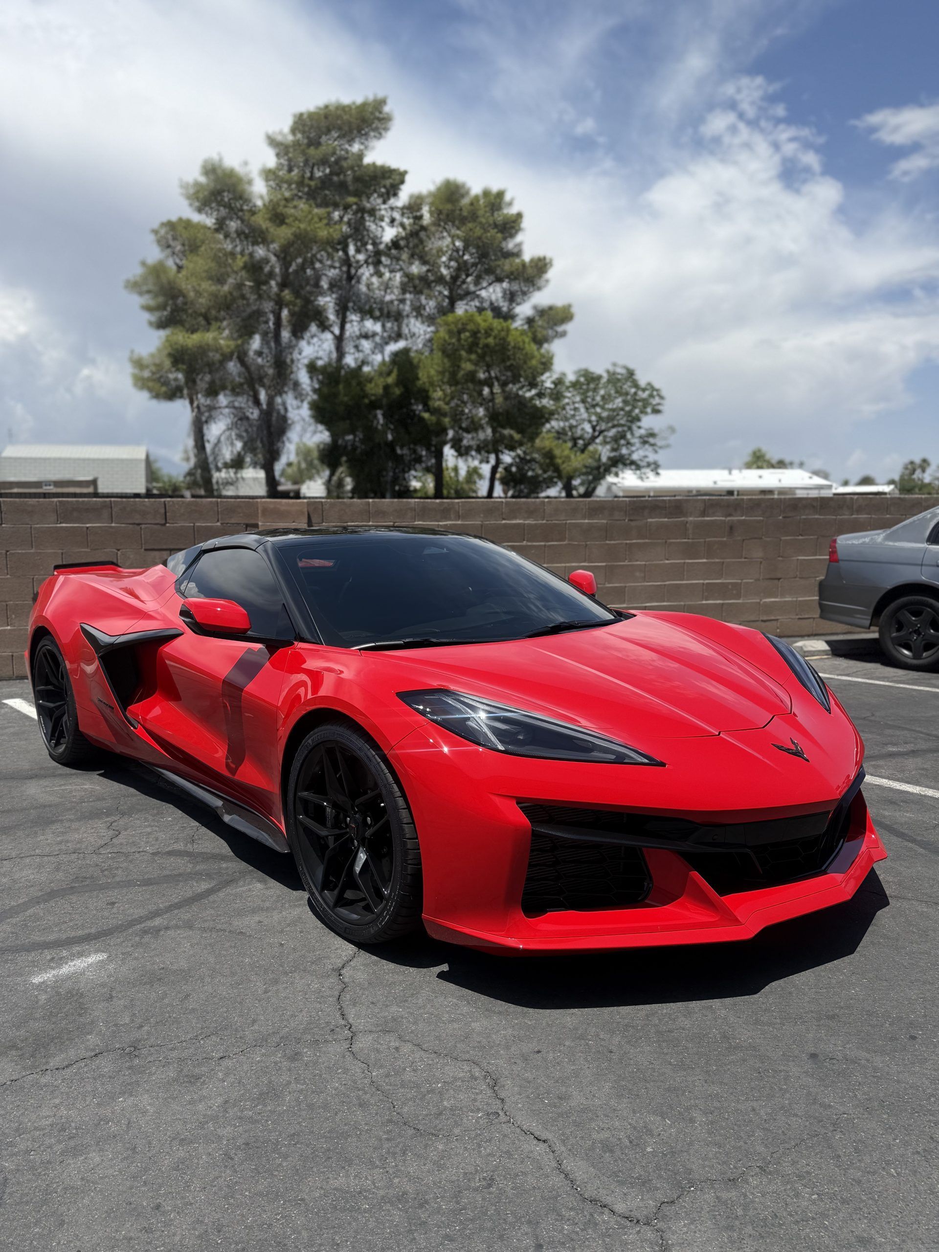 Red sports car parked on a sunlit asphalt lot with trees and a wall in the background