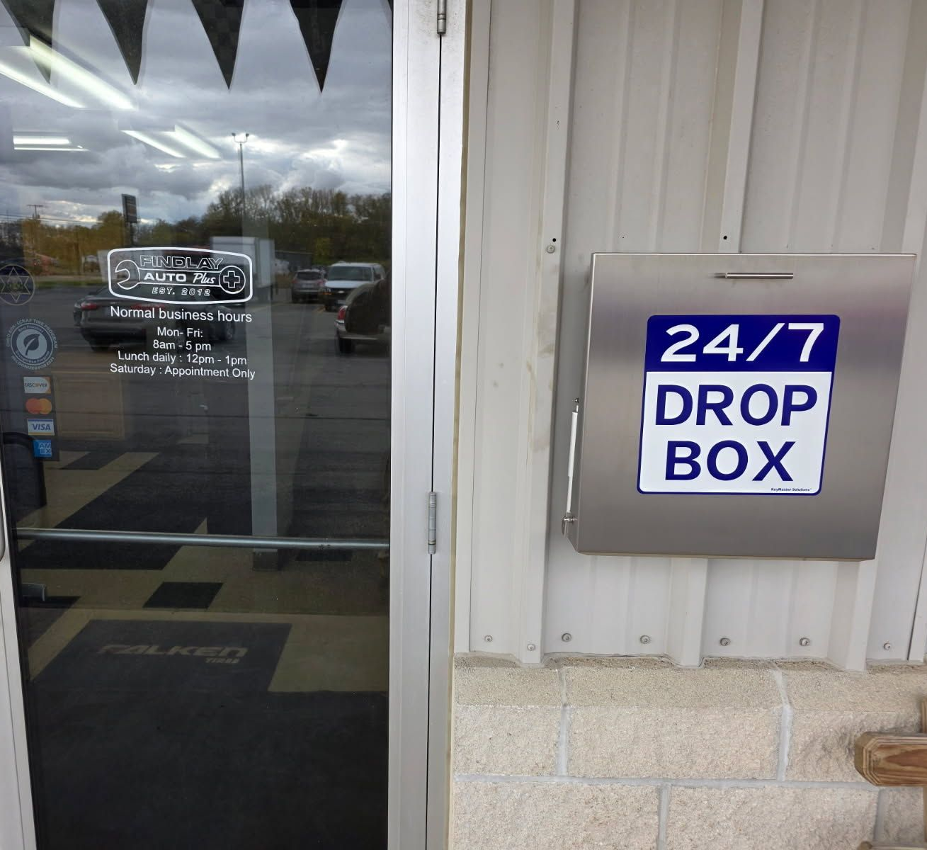 Silver drop box with 24/7 label on a building wall next to a glass door. | Findlay Auto Plus