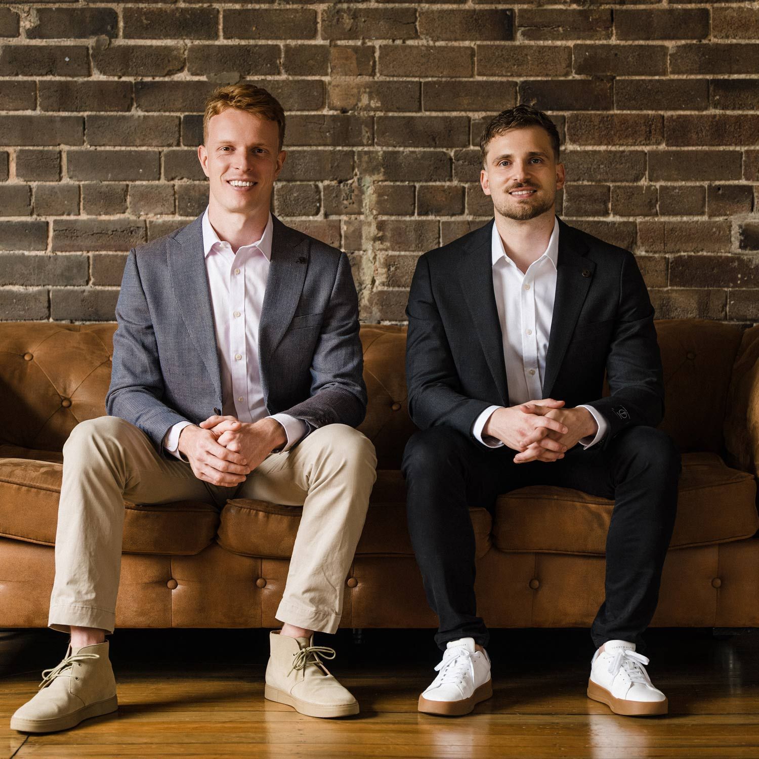 Two men in suits sit on a leather couch against a brick wall