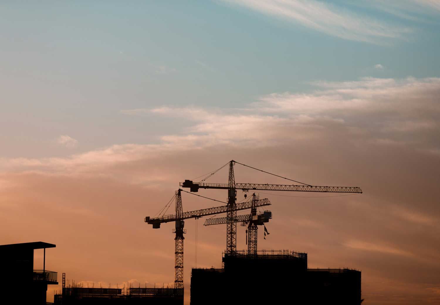 Construction cranes silhouetted against an orange and blue sunset sky