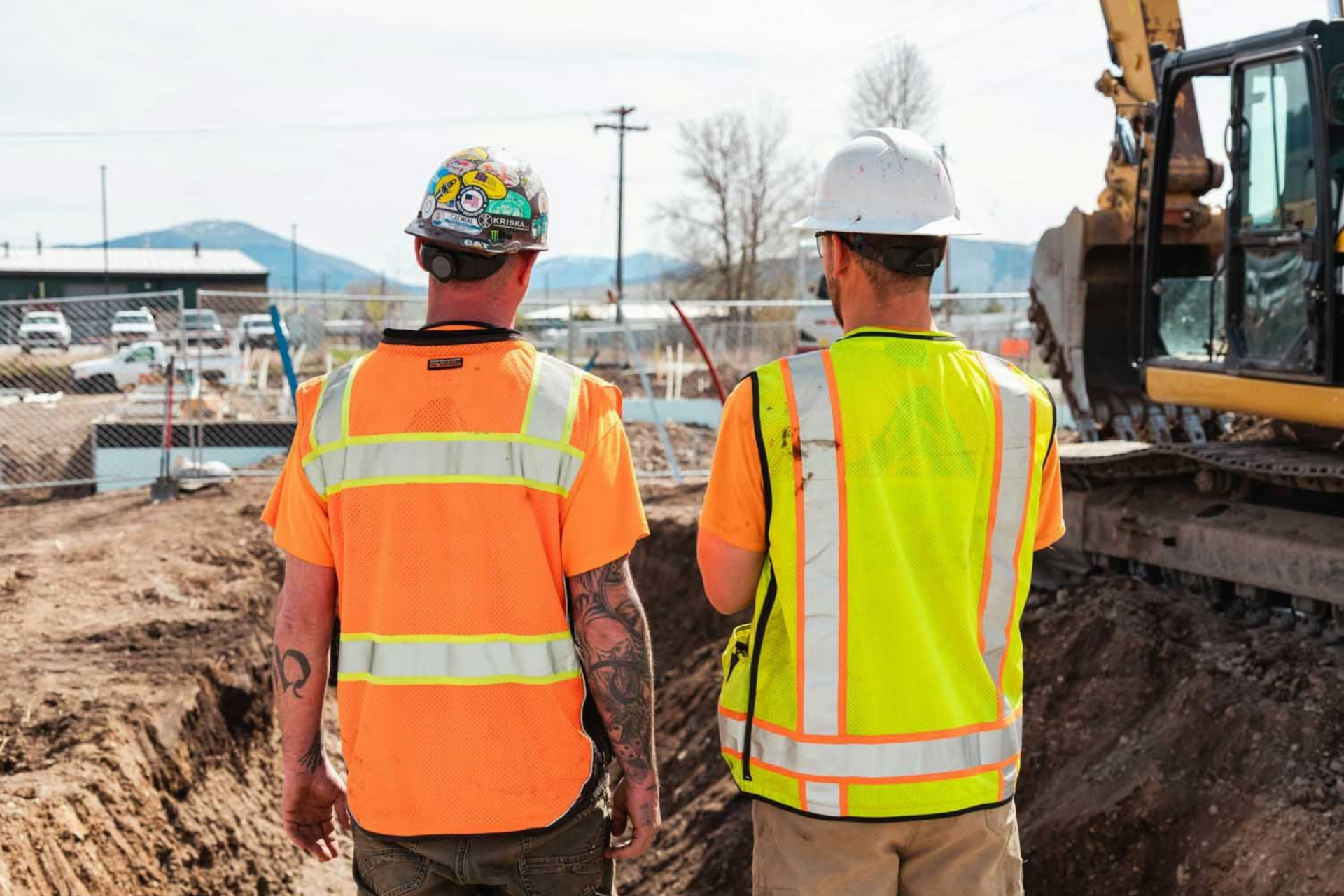 Two construction workers in safety vests stand near a trench at a construction site