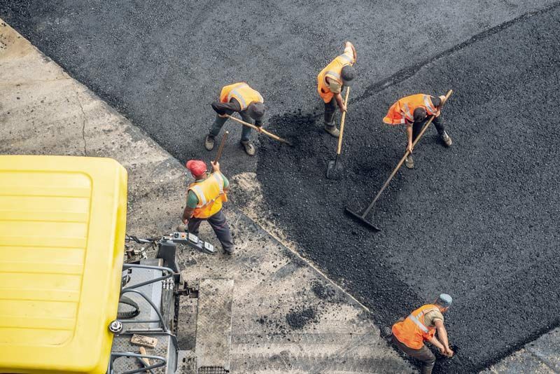 Road workers in orange vests and hard hats, raking asphalt next to a yellow truck