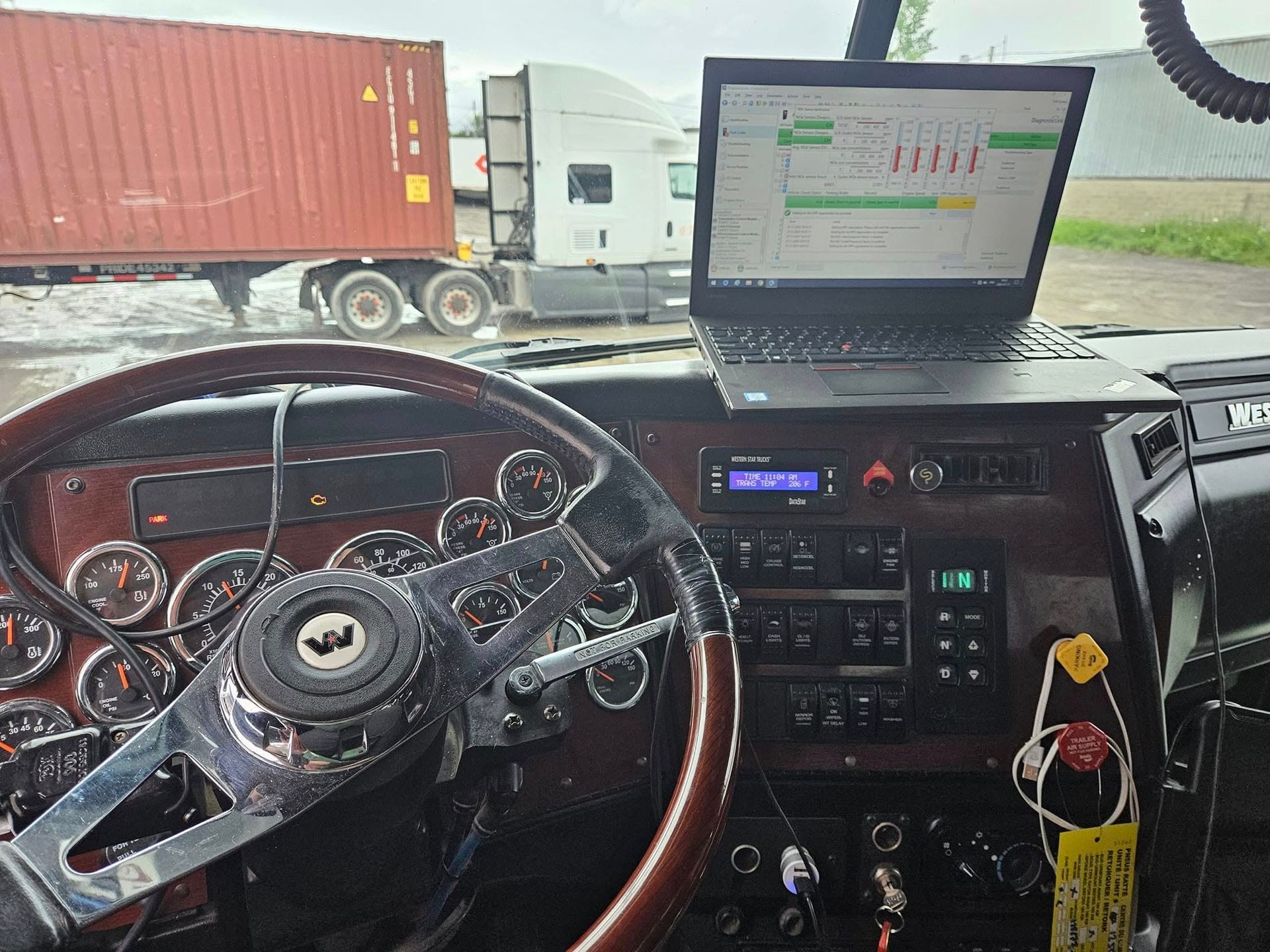 Vue intérieure d'une cabine de semi-remorque avec un ordinateur portable affichant des données ; un autre camion et un conteneur de fret sont visibles.