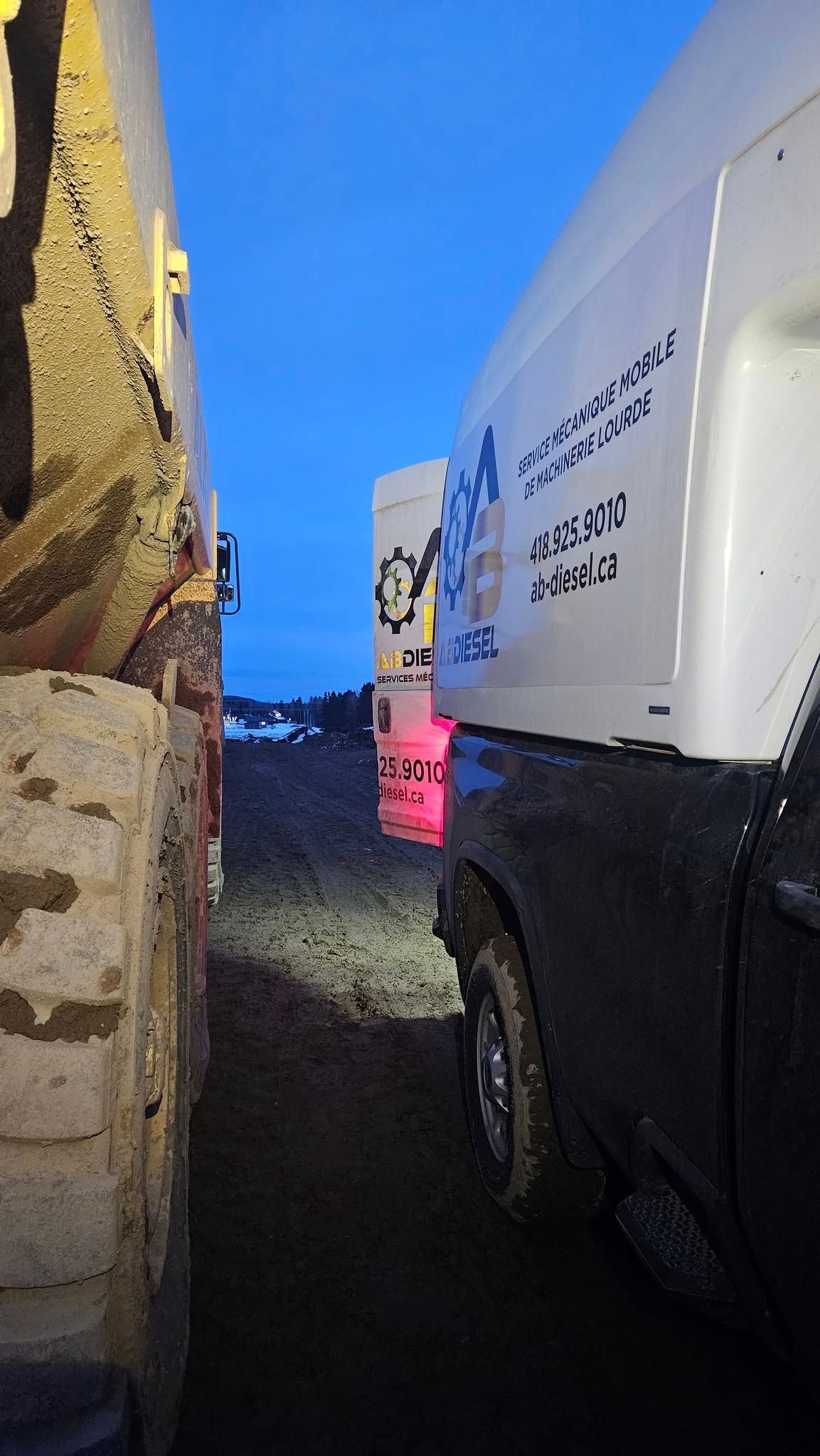 Camions garés les uns à côté des autres sur un chemin de terre au crépuscule. L'un est blanc avec du texte, l'autre est jaune.