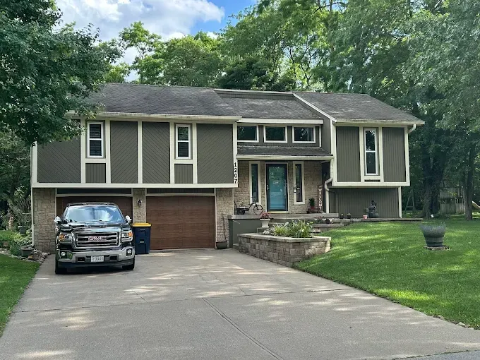 Two-story house with green siding, brown garage doors, and a gray car in the driveway. Landscaping includes a stone retaining wall and a manicured lawn.