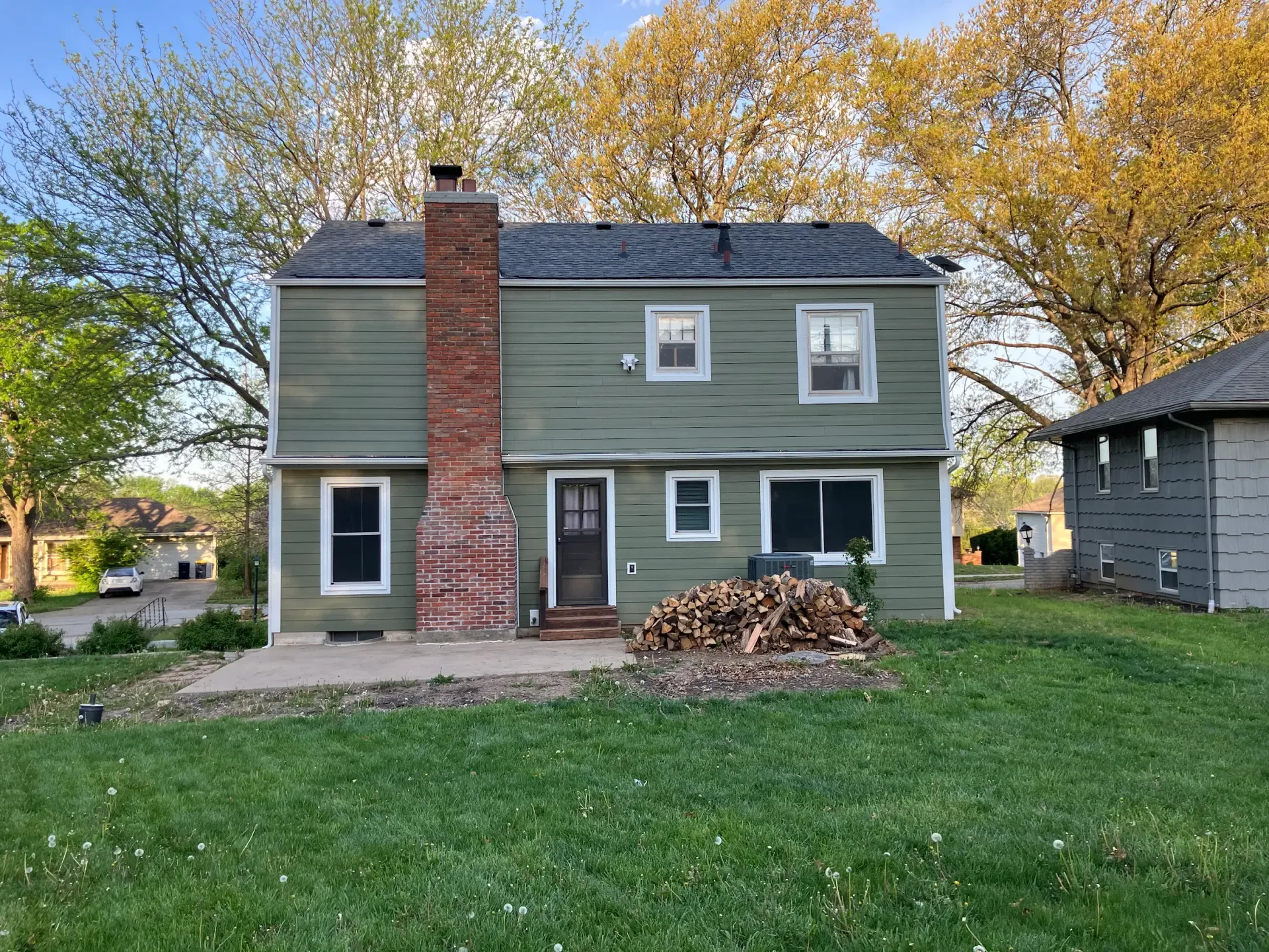Back of a two-story green house with a brick chimney. A stack of firewood sits near the back door on a stone patio.