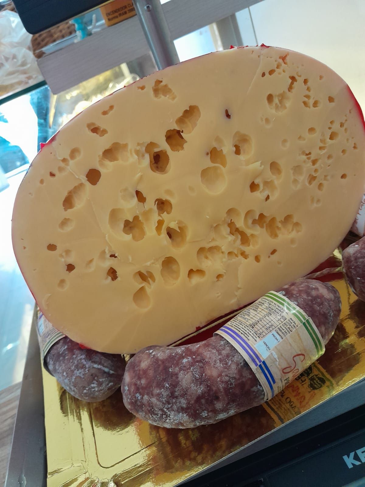 Queso grande, redondo y con agujeros, que reposa sobre una balanza junto a dos salchichas.