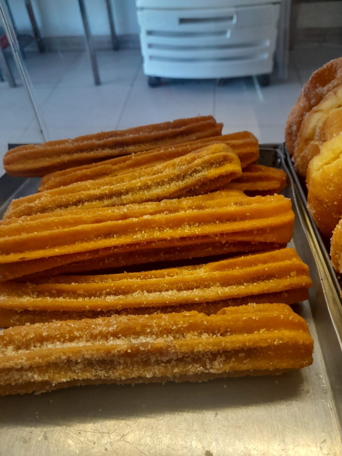 Una bandeja de churros dorados espolvoreados con azúcar, junto a una exposición de pasteles.