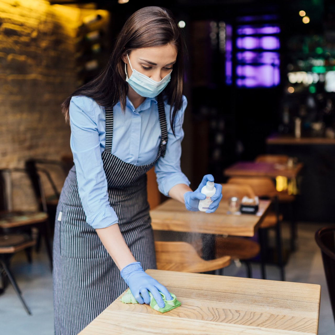 Young Caucasian female waitress wearing an apron, face mask and gloves, cleaning the tables using a wet wipe and a sanitizer