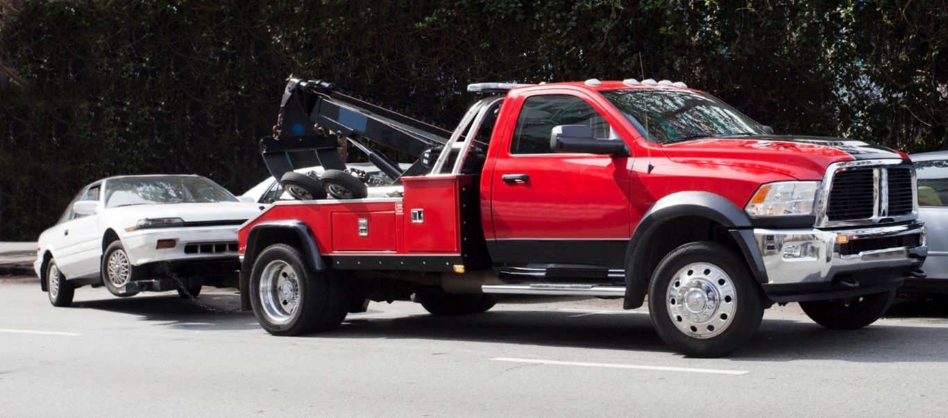 A red tow truck towing a white car on a street, with a backdrop of trees.