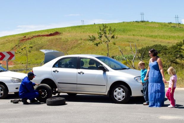 Mechanic changing a tire on a silver car; family watches roadside, sunny day.
