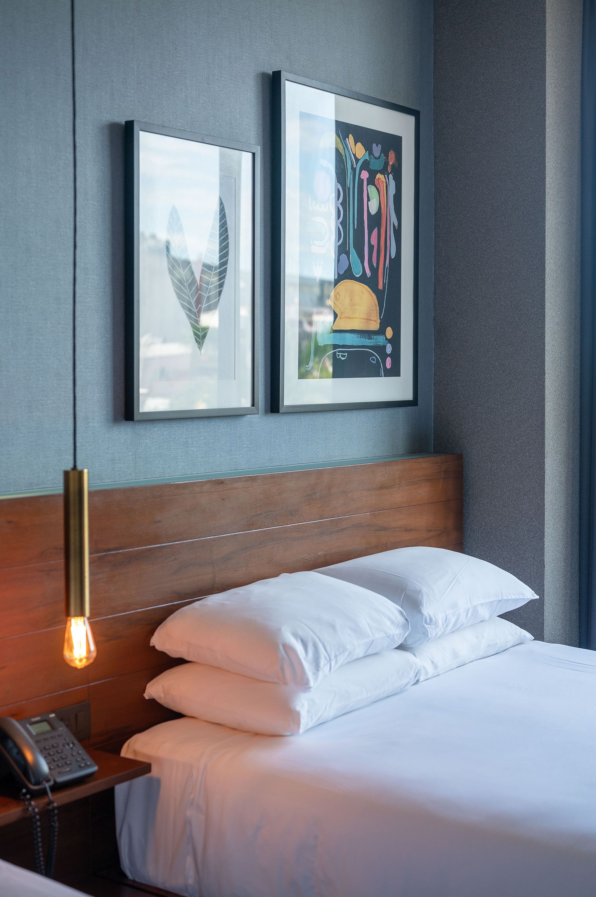 A hotel room with a bed , two paintings on the wall and a lamp hanging from the ceiling.