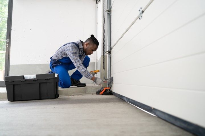 Worker in blue overalls kneeling to repair a garage door track with tools nearby