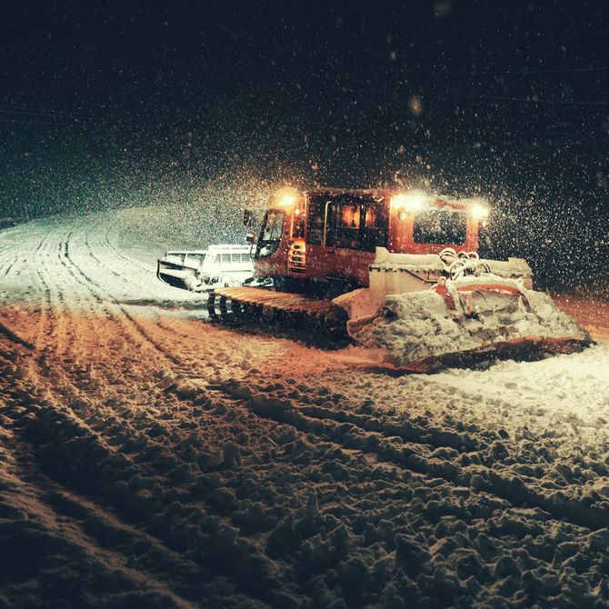 Snowplow clearing a snowy road at night with bright headlights and falling snow