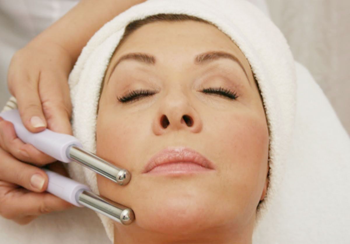 Woman receiving facial treatment with hand-held devices, eyes closed, wrapped in a white towel.