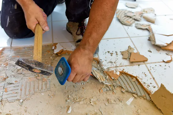 A man is using a hammer to remove tile from the floor.