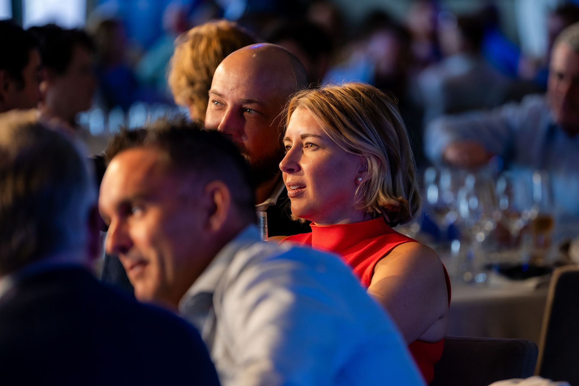 People at a dinner event, a woman in a red top and a man look toward a center focal point.
