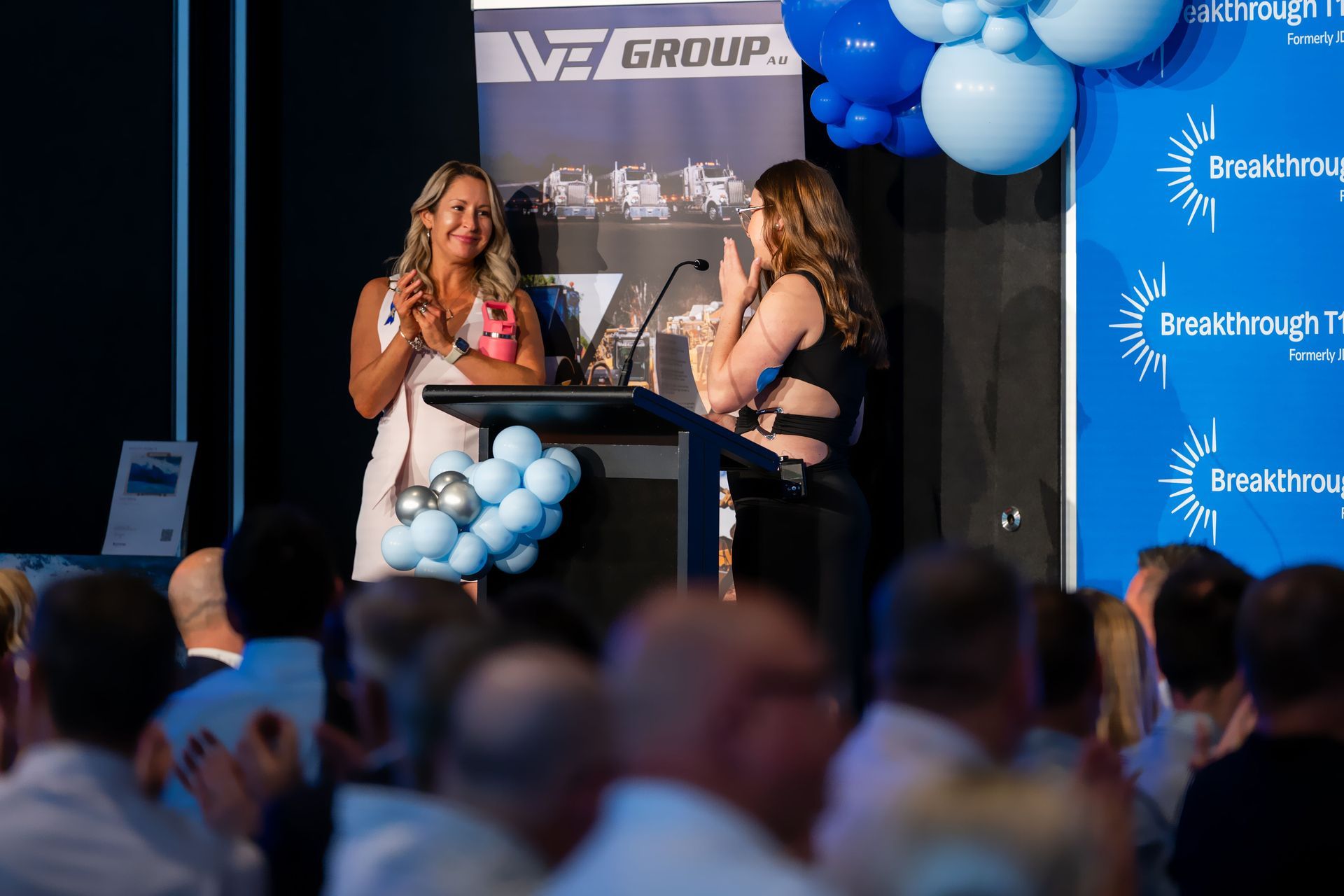 Two women on stage at event, one speaking at podium, others applauding in background.