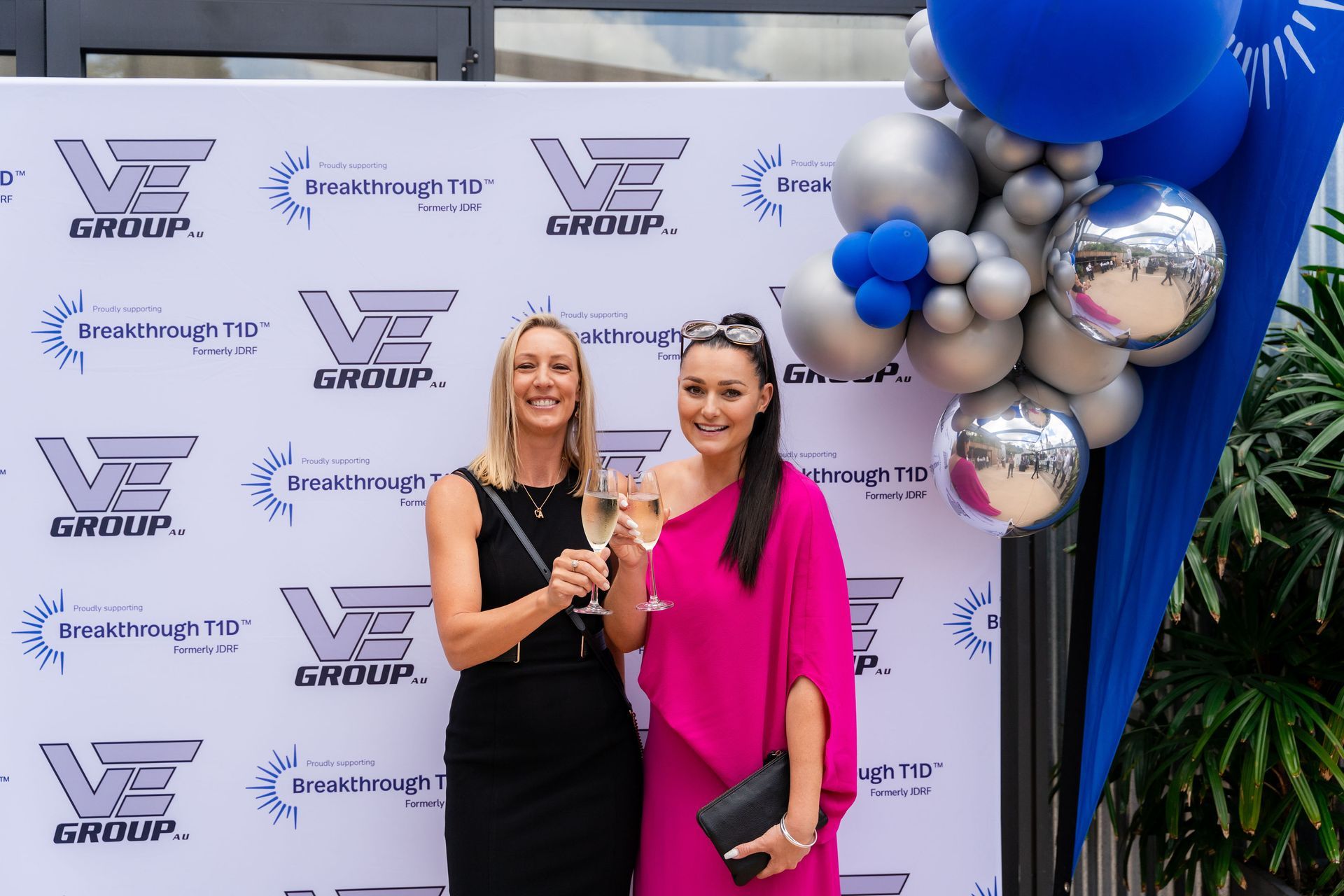 Two women toasting with champagne flutes at a VE Group event with branded backdrop and balloons.