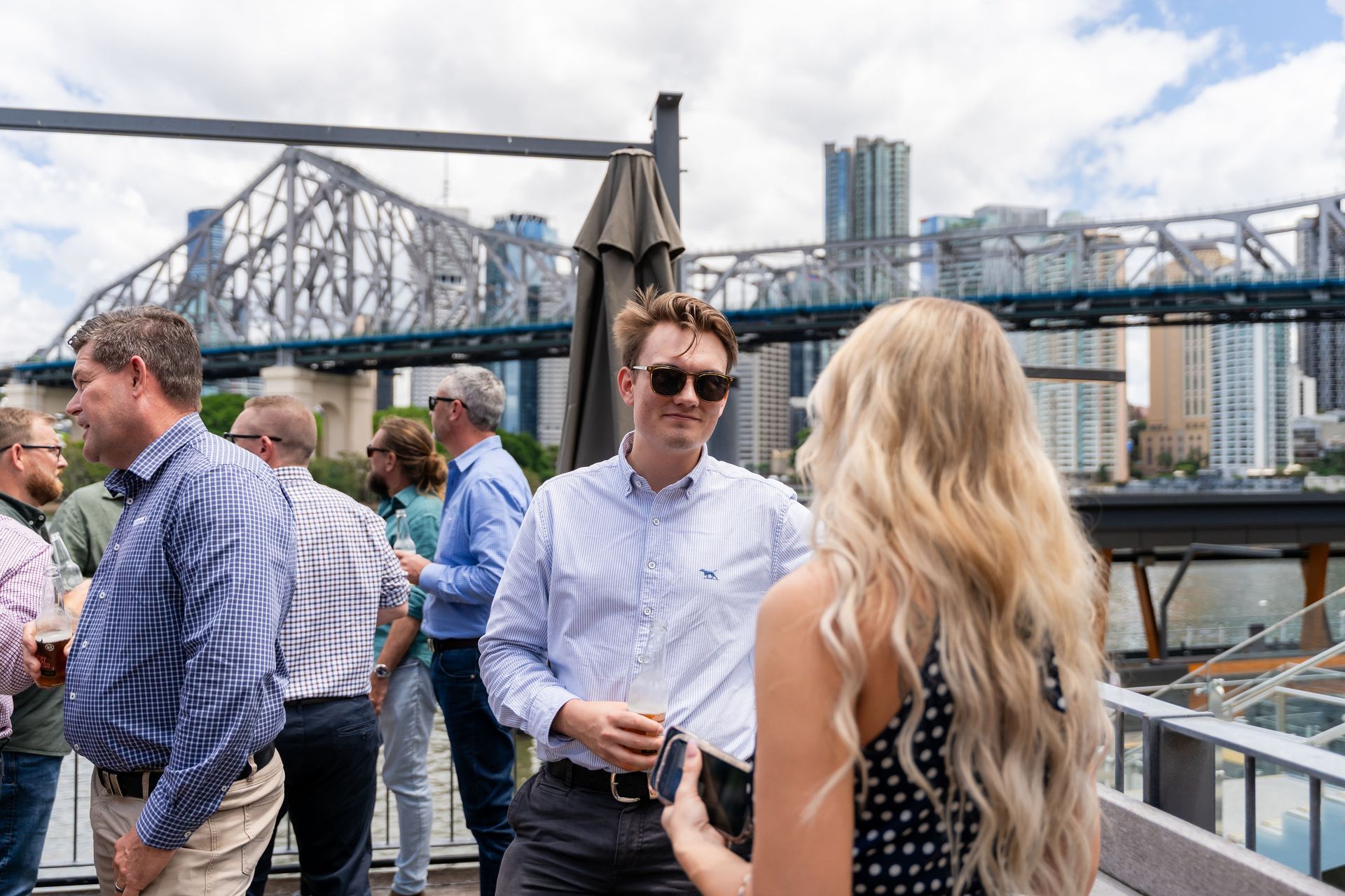 People socializing on a rooftop, Brisbane, with a bridge and city skyline in the background.