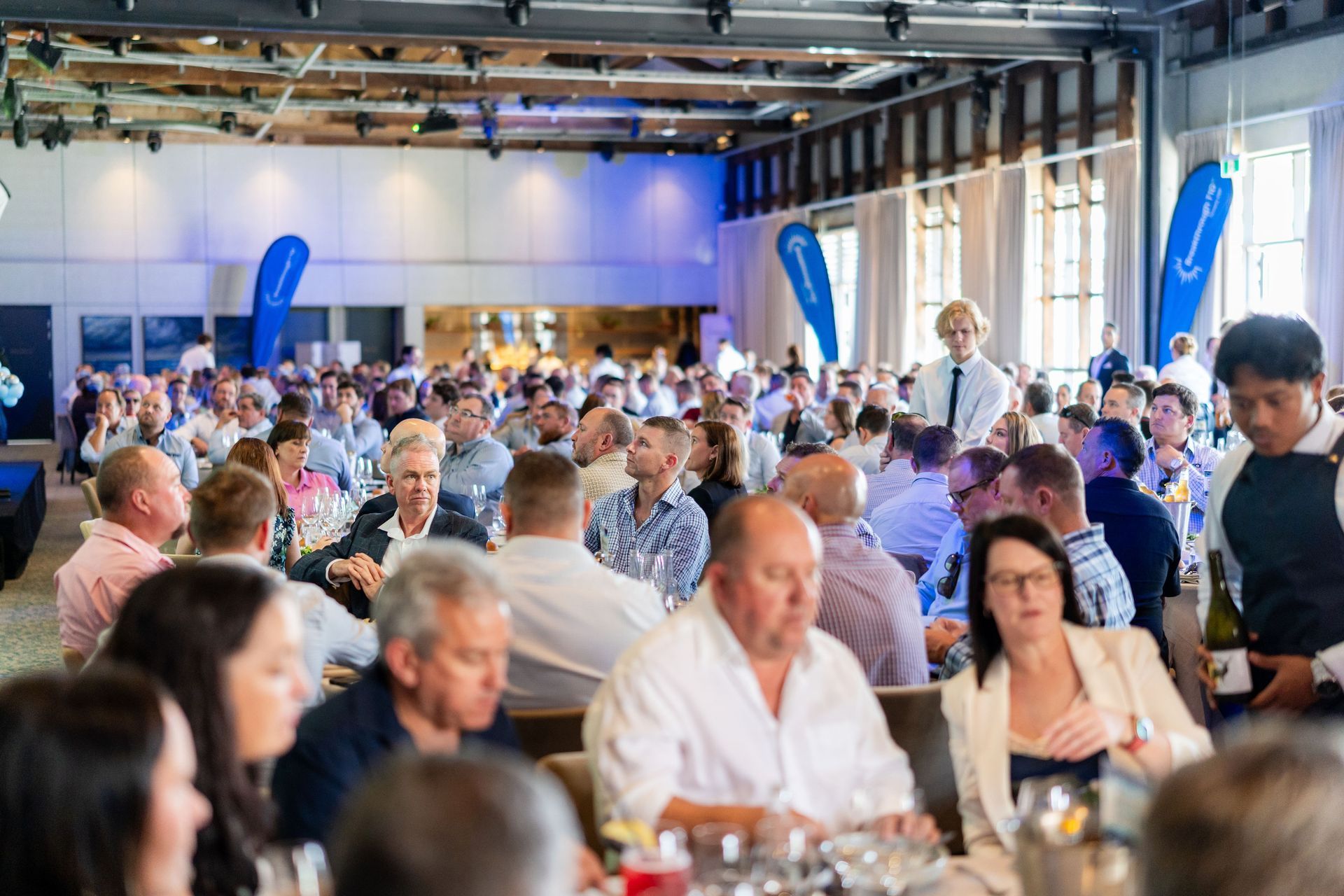 Large event: crowd seated at tables in a bright room, staff serving, blue accents.