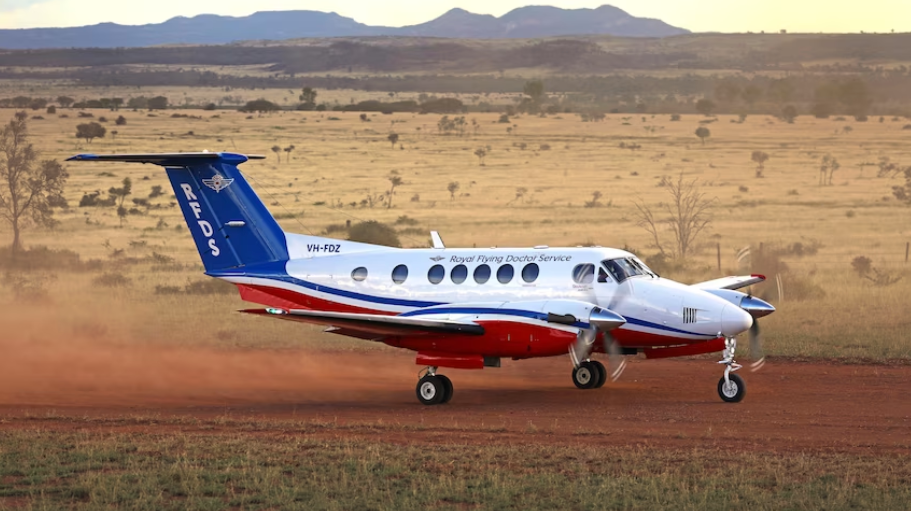 Small airplane with white, blue, and red paint, kicking up dust on a dirt runway with a desert landscape in the background.