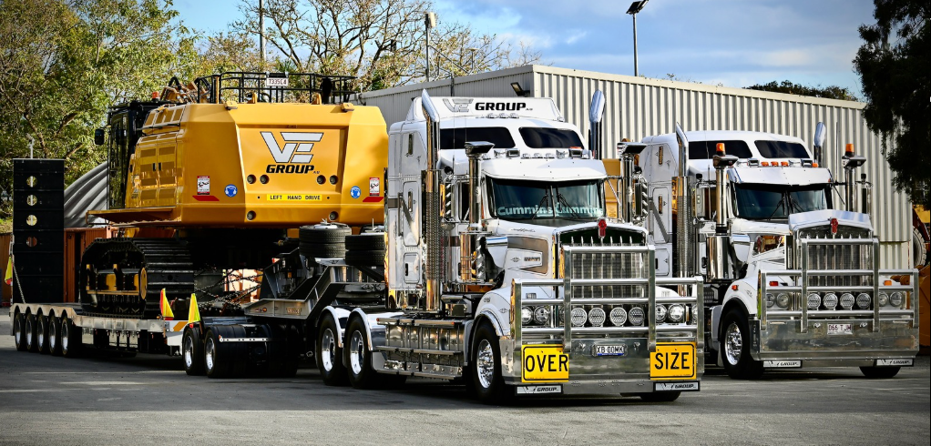 Two white semi trucks with chrome accents; a large yellow machine is on a trailer hitched to the first truck.