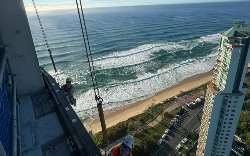 Workers on high-rise building with ocean view, cables, and a nearby building.