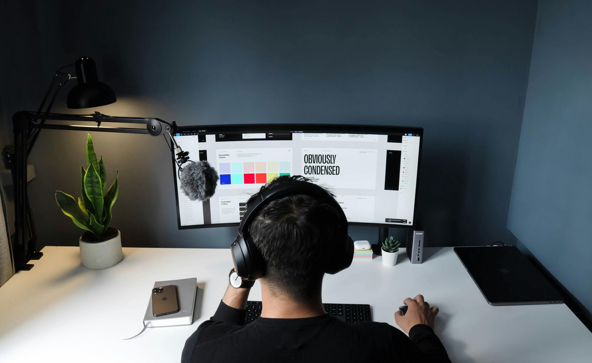 A person wearing headphones works at a desk with a curved monitor, plant, and desk lamp in a minimalist workspace.