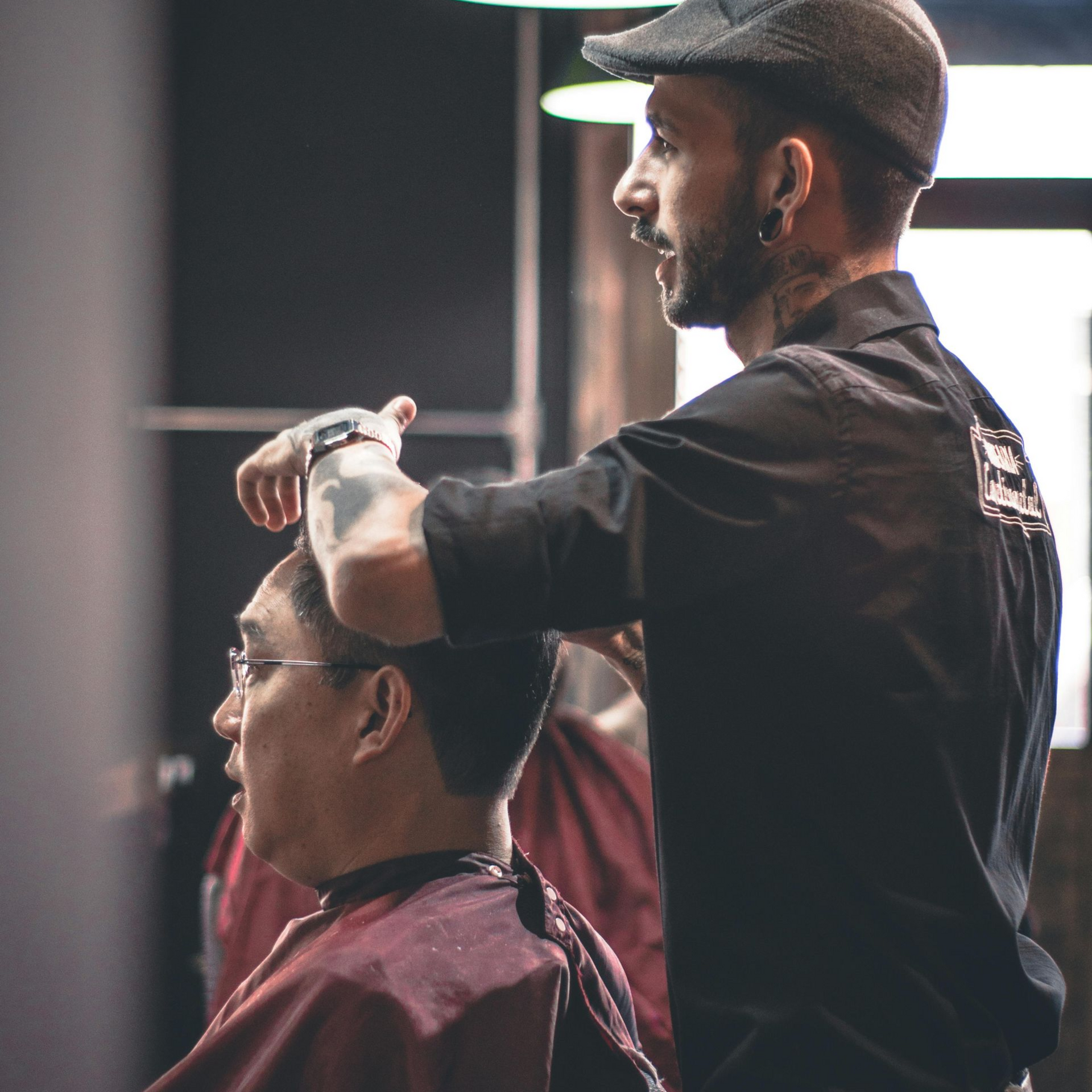 Barber in black shirt and cap cutting client's hair in a barbershop.