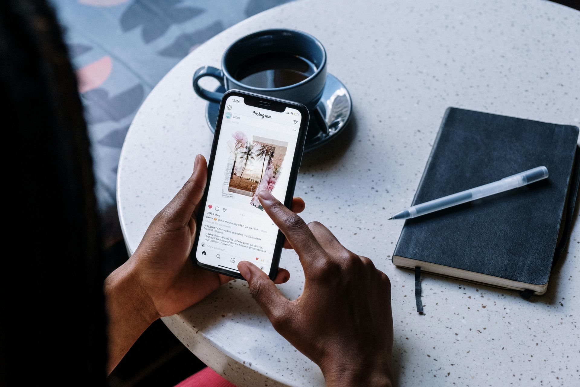 Person using a smartphone on a table, next to a coffee cup and a notebook with a pen.