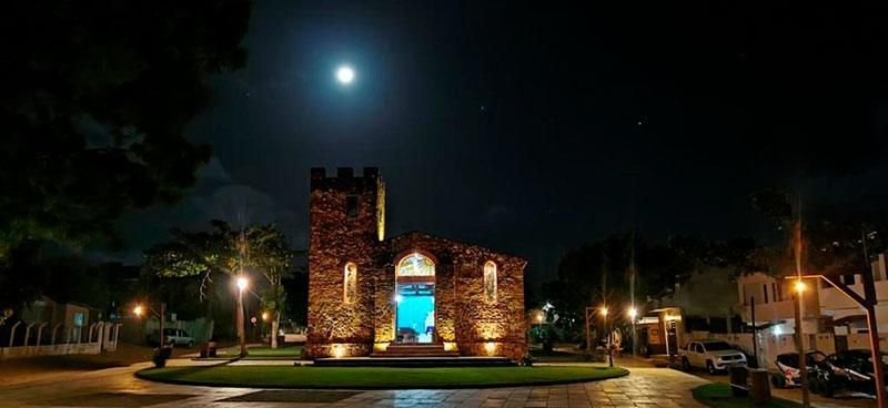 Uma igreja de pedra iluminada à noite sob a luz da lua cheia, rodeada por um gramado e iluminada por lâmpadas externas de luz quente.