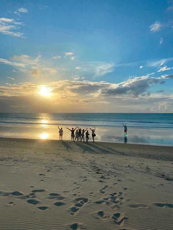 Um grupo de pessoas em pé numa praia ao pôr do sol, com os braços erguidos, refletindo-se na areia molhada junto ao oceano.
