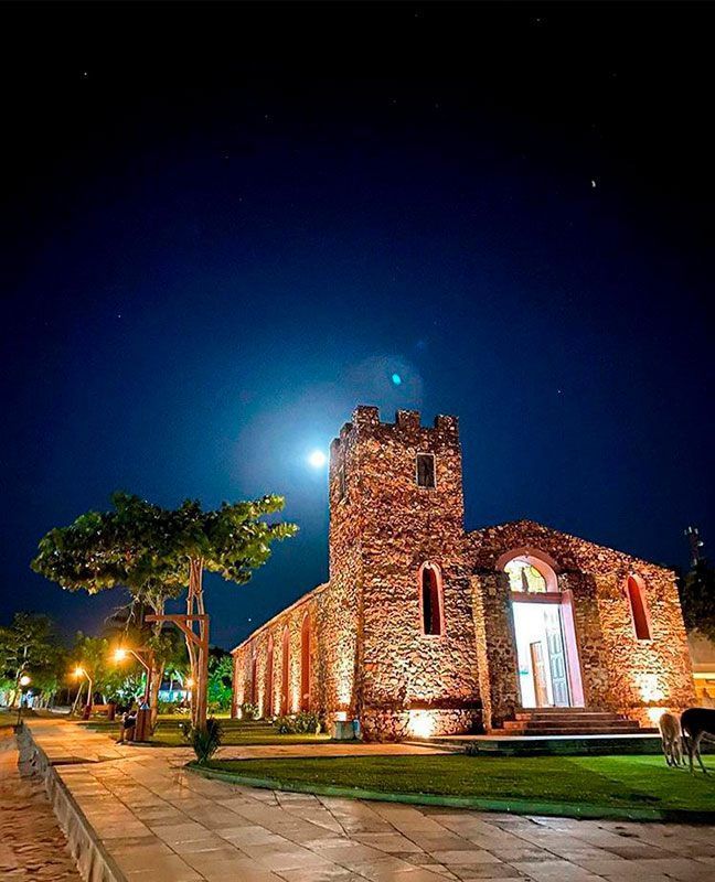 Uma igreja de pedra iluminada à noite sob a luz da lua cheia, com um caminho pavimentado e árvores em primeiro plano.