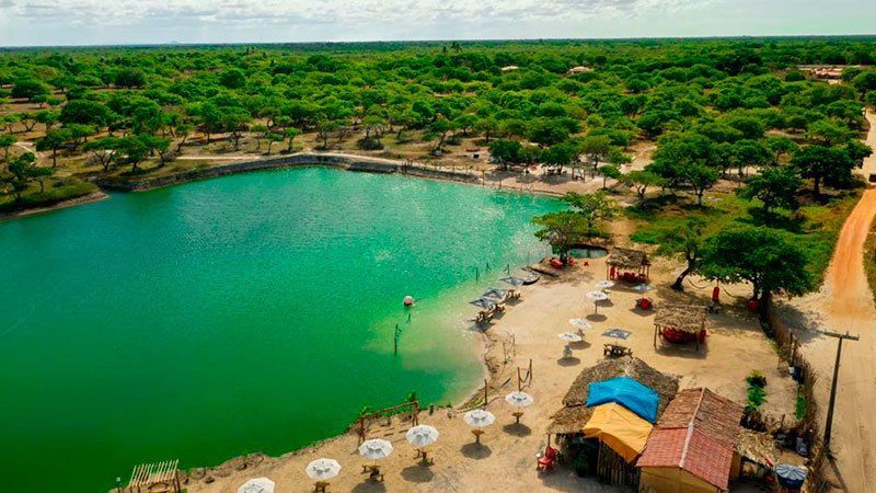 Vista aérea de uma lagoa de cor turquesa brilhante rodeada por árvores, com guarda-sóis e pequenas estruturas na areia.