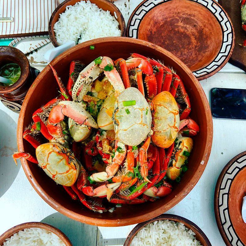Uma grande tigela de terracota cheia de caranguejos cozidos, guarnecida com cebolinha verde, acompanhada de pequenas tigelas de arroz branco.