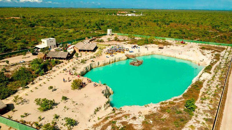 Vista aérea de uma lagoa de um azul turquesa vibrante, rodeada por margens arenosas, pequenas construções e uma exuberante floresta verde.