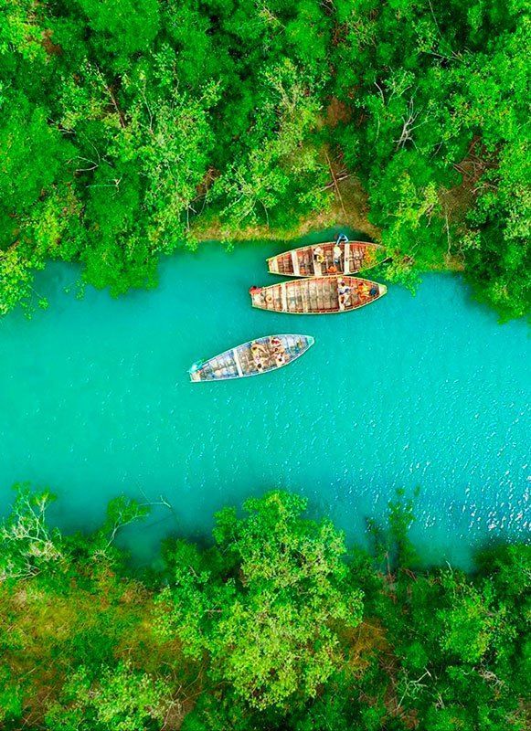 Vista aérea de três barcos de madeira desgastados flutuando em um rio de águas turquesa vibrantes, cercado por uma densa floresta verde.