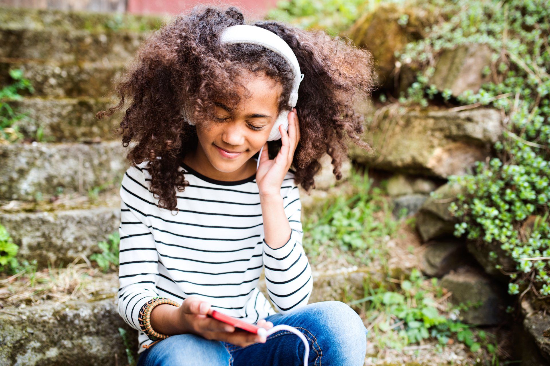 A young girl is sitting on a set of stairs listening to music on her phone.