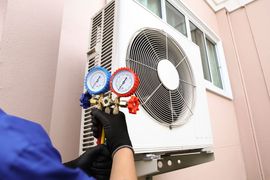A technician wearing black gloves uses a manifold gauge set to service an outdoor air conditioning unit.